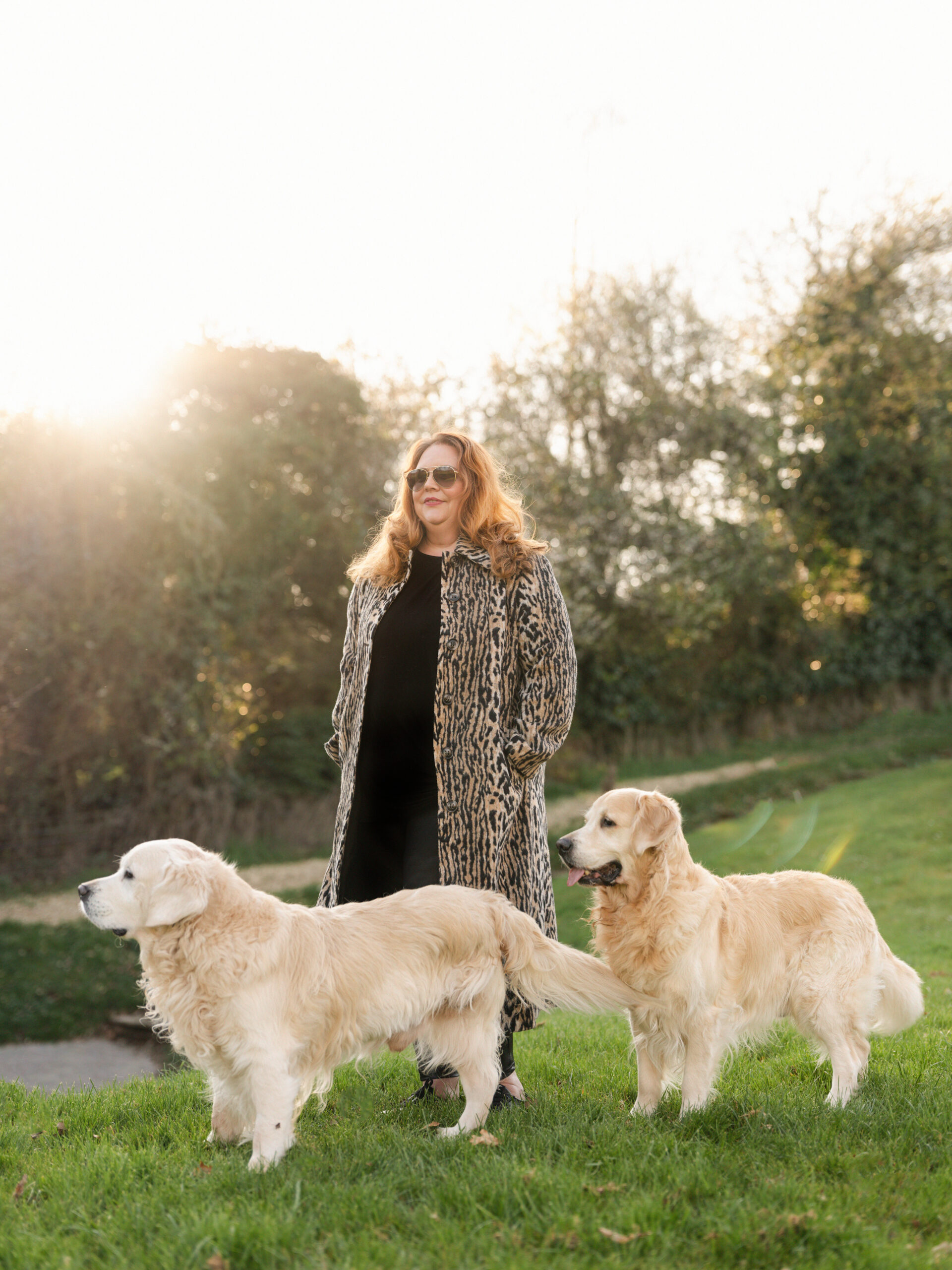 Woman in a leopard-print coat and sunglasses standing in a sunlit park with two Golden Retrievers nearby